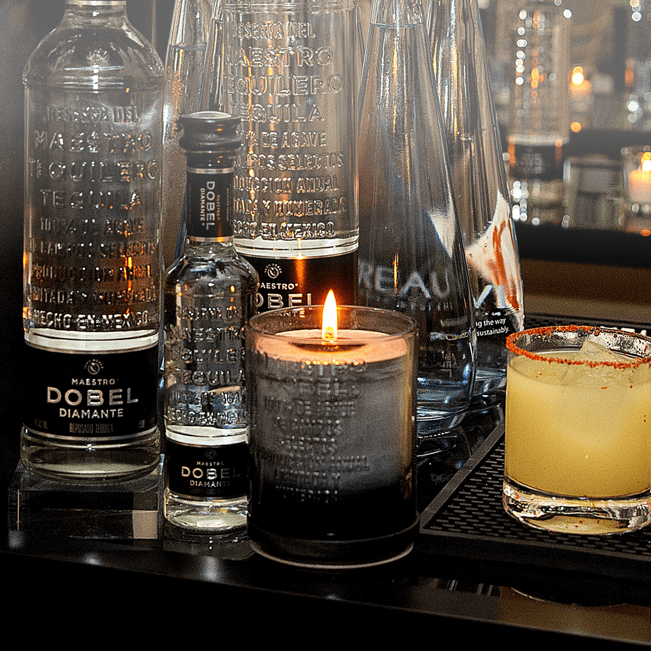 Dobel Cooked Agave Scented Candle lit on a table with Dobel Tequila bottles.
