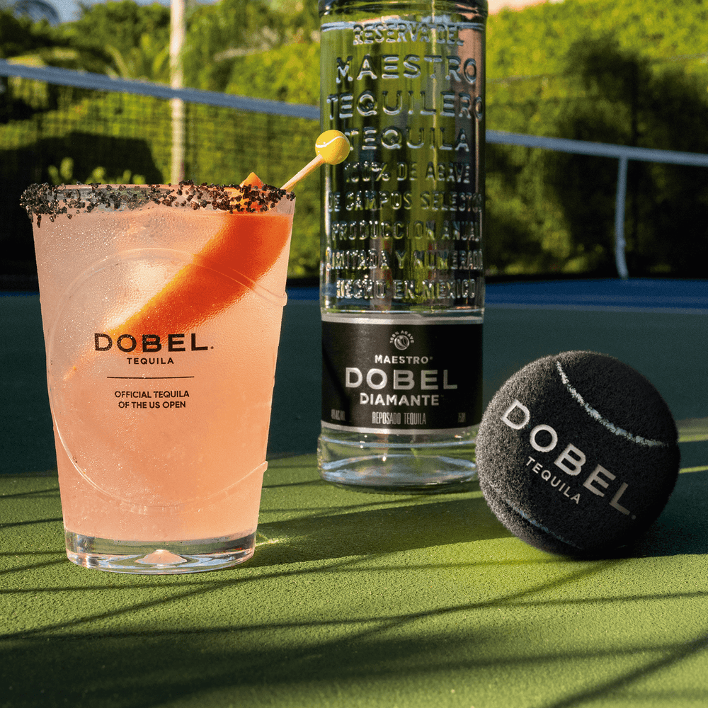 Black Dobel tennis ball with Ace Paloma cocktail and Dobel Diamante Cristalino bottle on a tennis court.