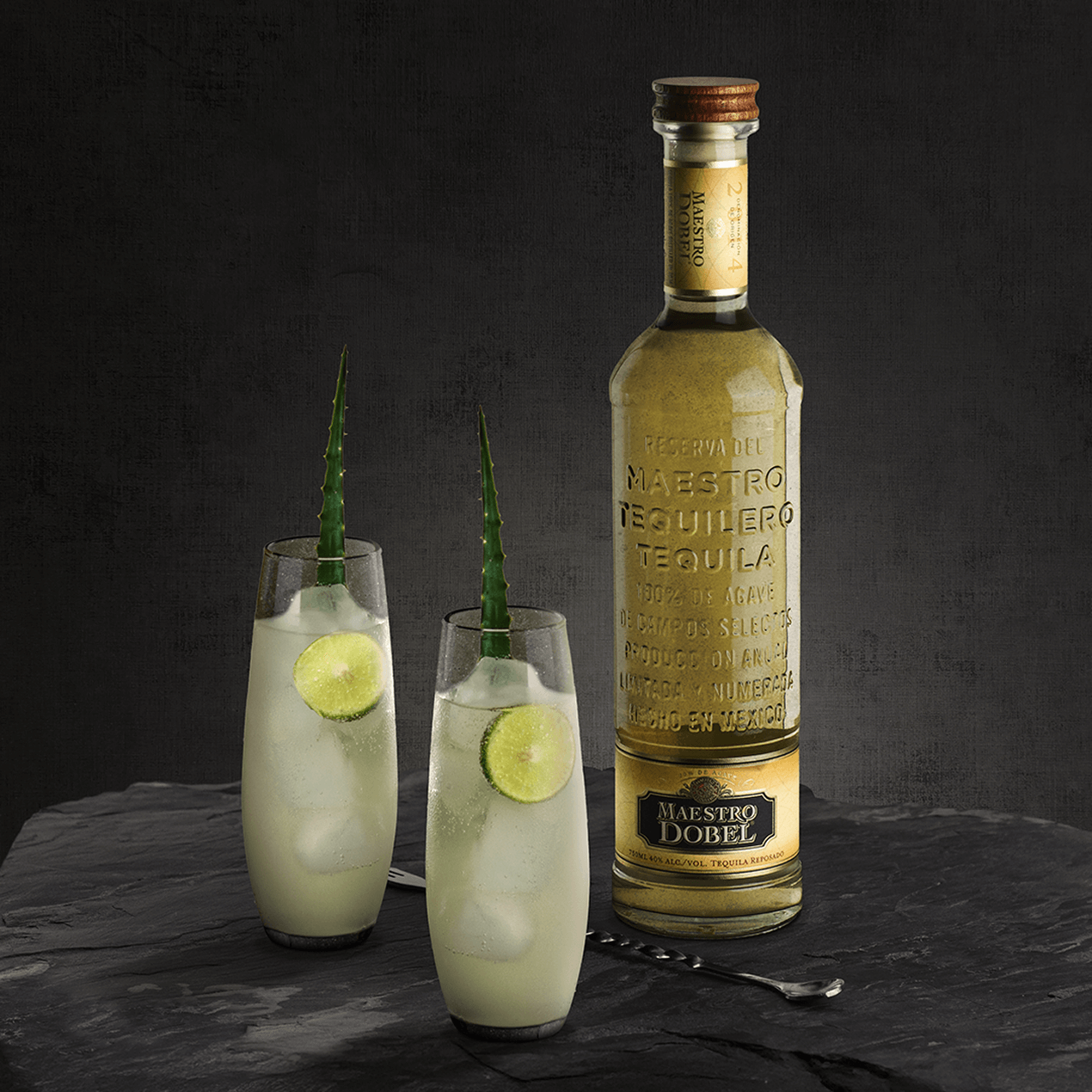 Dobel Reposado bottle with two lime-garnished cocktails on black surface