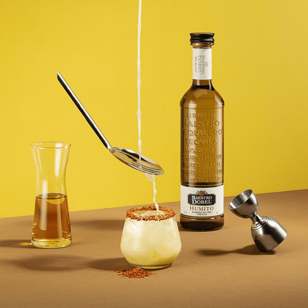 Dobel Humito bottle on a brown surface with a cocktail being poured through a spoon, surrounded by a jigger and a glass with chili rim.