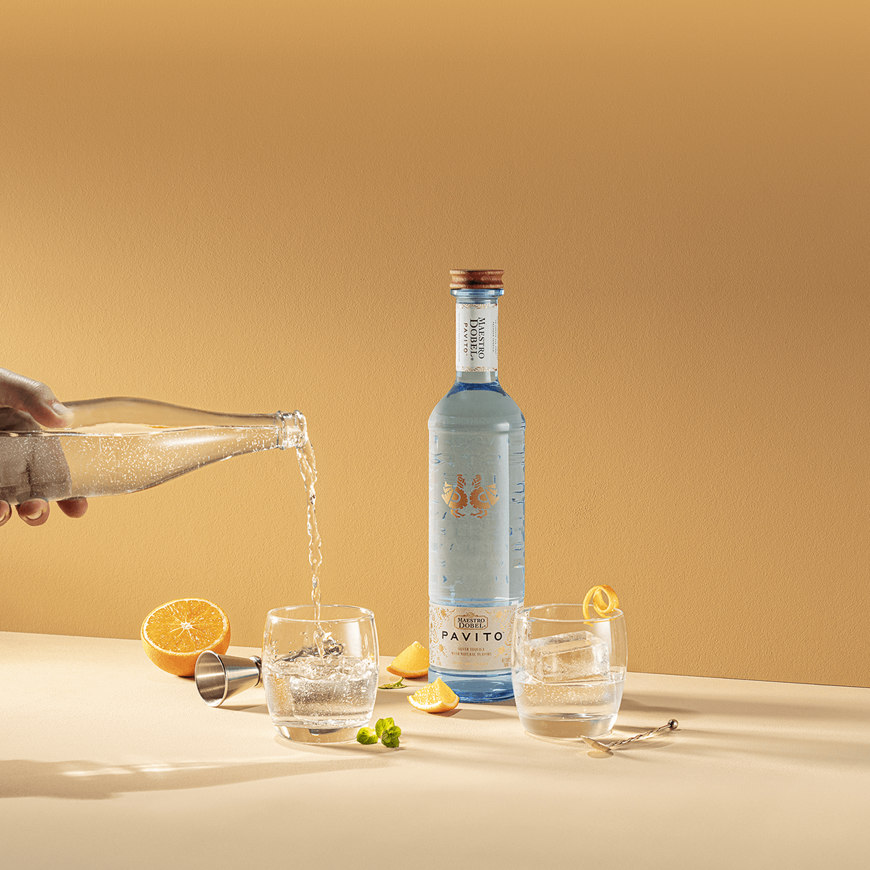 Dobel Pavito bottle on a table with two cocktail glasses, citrus garnish, and a hand pouring from a soda bottle.