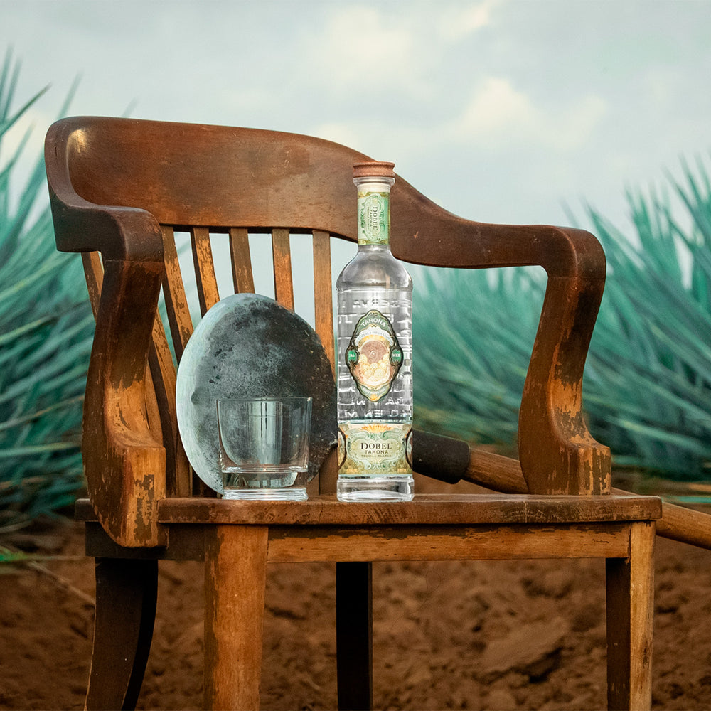 Dobel Tahona on a rustic wooden chair amid agave plants.