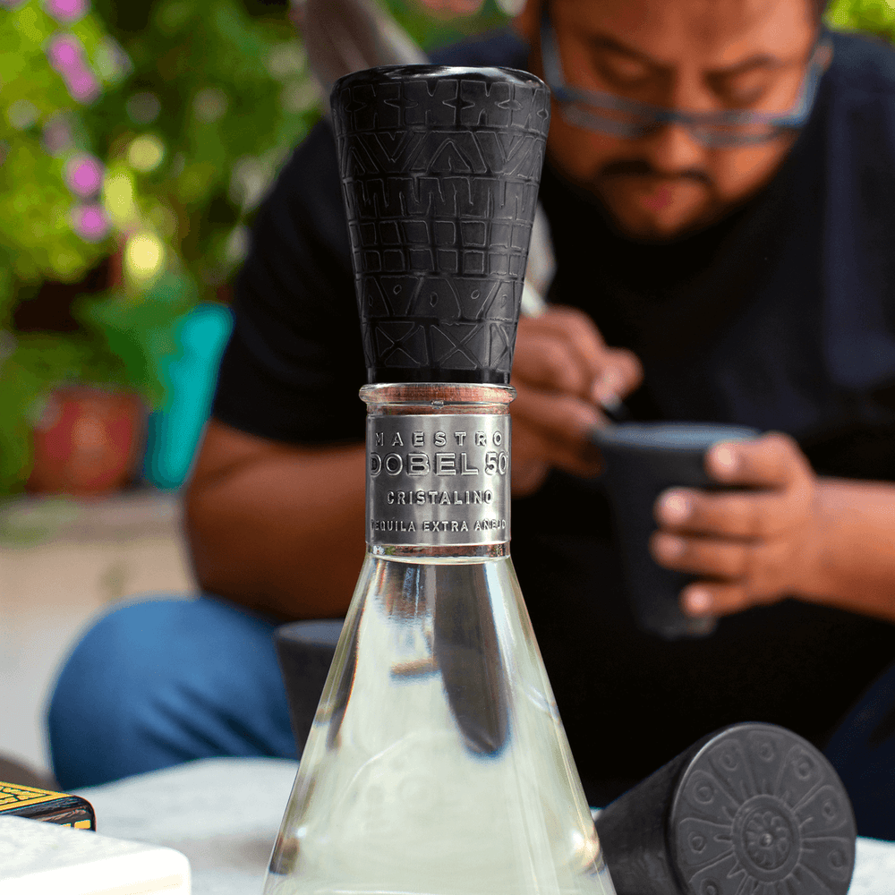 Dobel 50 Cristalino Onora Laca de Olinala bottle with a person crafting in the background.