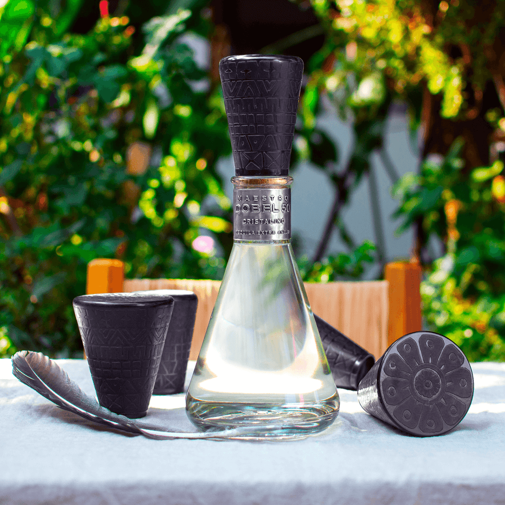 Dobel 50 Cristalino Onora Laca de Olinala bottle with artistic cups on a table outdoors.