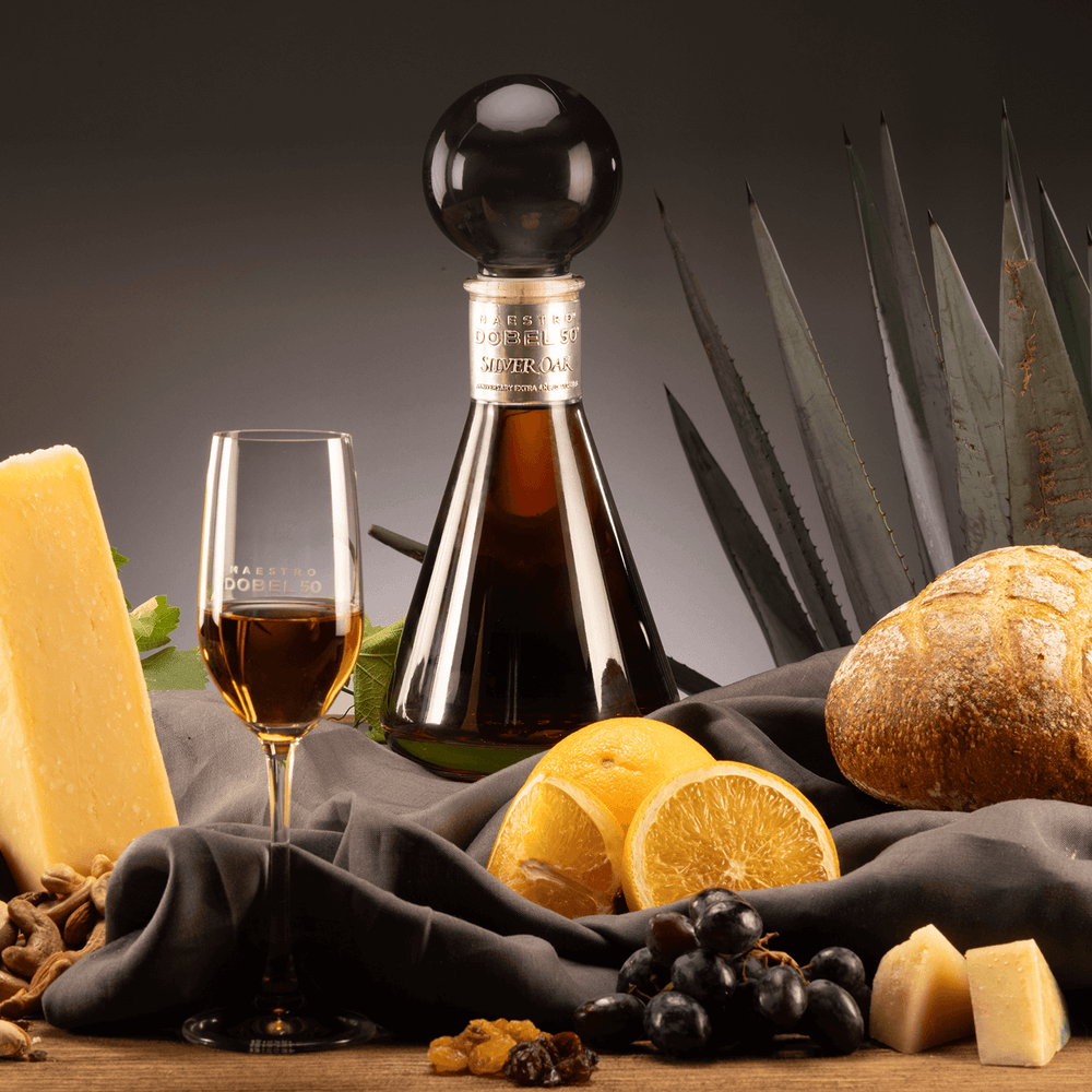 Dobel 50 Extra Añejo Silver Oak bottle with flute of poured tequila on a table with bread, cheese, and other snacks.