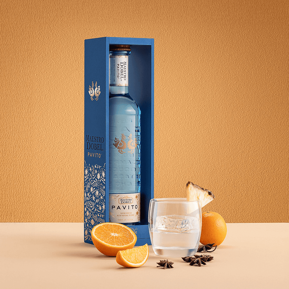 Dobel Pavito bottle in a blue gift box next to a glass of tequila and slices of orange and pineapple.