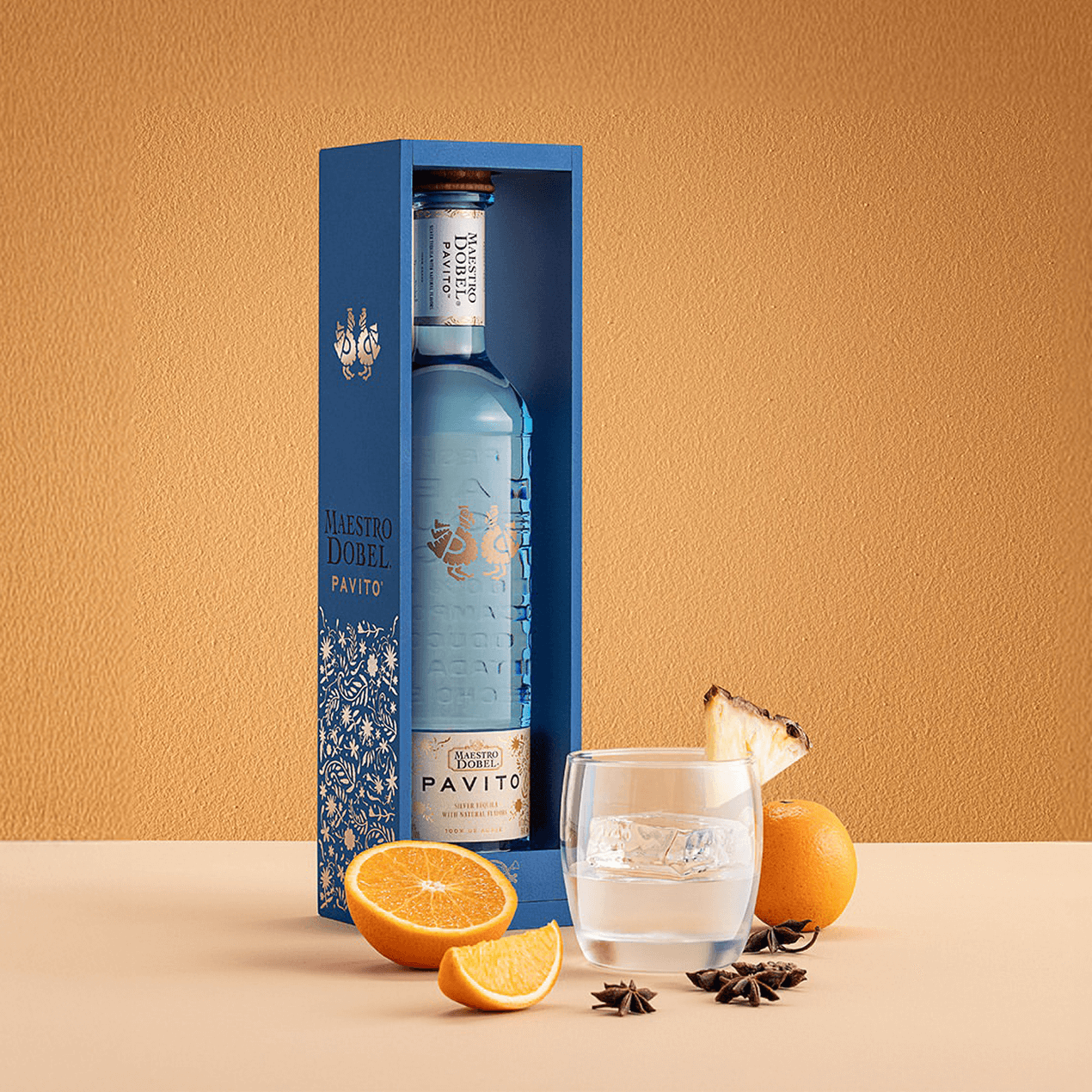 Dobel Pavito bottle in a blue gift box next to a glass of tequila and slices of orange and pineapple.